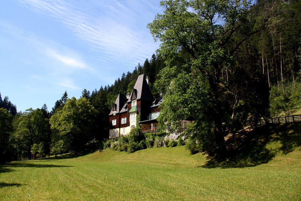 Ein imposantes Haus mit mehreren Türmen und grünem Dach, umgeben von Bäumen. Im Hintergrund ist eine sanfte Hügellandschaft sichtbar. | © nicoleseiser.at