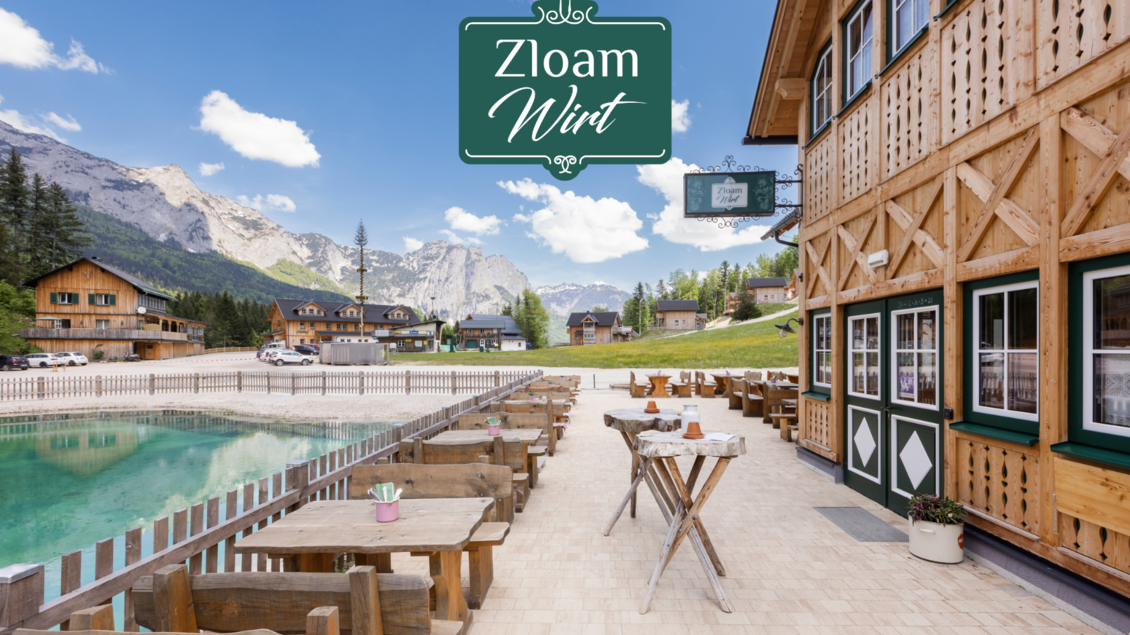 Ein malerisches Bergdorf mit Holzchalets und einer einladenden Terrasse. Im Vordergrund sieht man einen klaren See und schöne Berglandschaften im Hintergrund. | © www.zloam.at