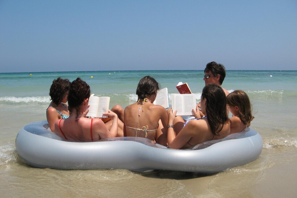 Eine Gruppe von Menschen sitzt auf einem aufblasbaren Schwimmring im Wasser. Alle lesen Bücher am Strand unter klarem Himmel. | © (c)Margreid Tiefenbrunner