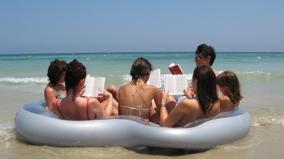 Eine Gruppe von Menschen sitzt auf einem aufblasbaren Schwimmring im Wasser. Alle lesen Bücher am Strand unter klarem Himmel. | © (c)Margreid Tiefenbrunner