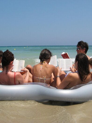 Eine Gruppe von Menschen sitzt auf einem aufblasbaren Schwimmring im Wasser. Alle lesen Bücher am Strand unter klarem Himmel. | © (c)Margreid Tiefenbrunner
