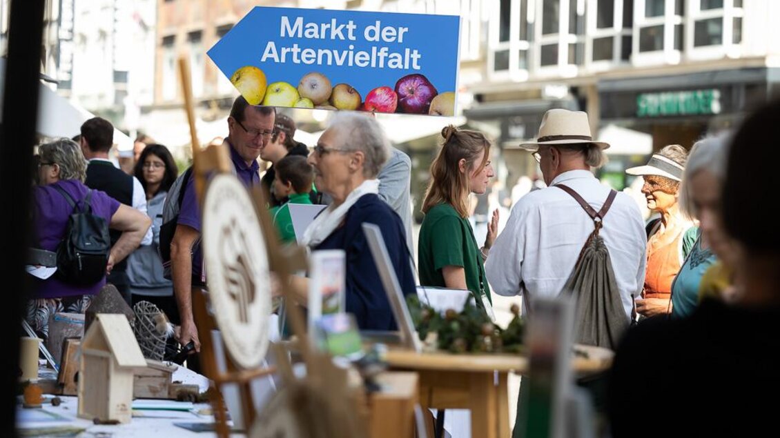 Ein belebter Markt mit vielen Besuchern und Ständen. Ein blaues Schild mit der Aufschrift „Markt der Artenvielfalt“ ist im Vordergrund sichtbar. | © c_Oliver_Wolf.