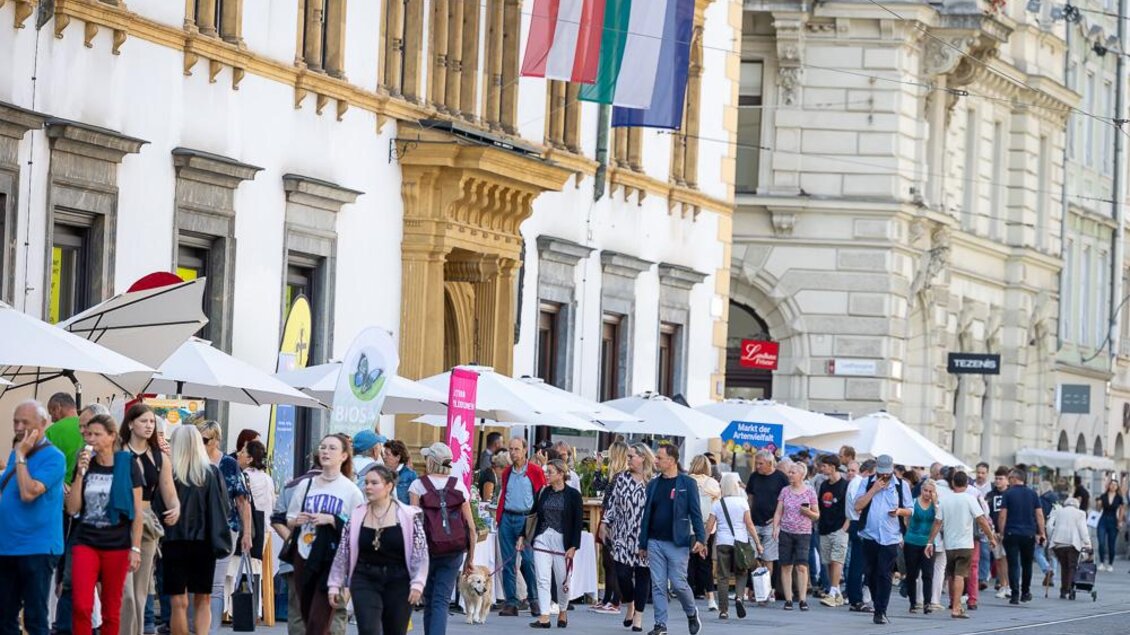 Eine belebte Straße mit vielen Menschen, die entlang von Ständen spazieren. Über den Ständen hängen Sonnenschirme und die Atmosphäre wirkt freundlich und einladend. | © _c_Oliver_Wolf