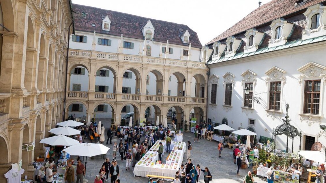 Ein belebter Innenhof mit vielen Menschen und weißen Sonnenschirmen. In der Mitte steht ein langer Tisch mit verschiedenen Ständen und Verkaufsständen. | © _c_Oliver_Wolf