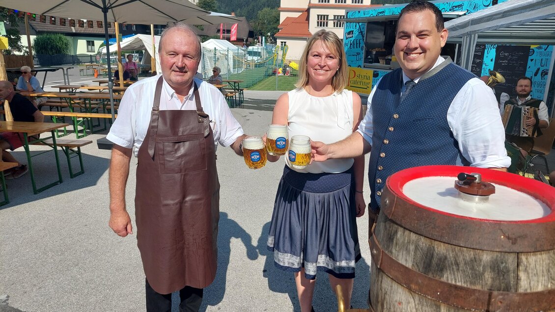 Drei Personen lächeln und halten Bierkrüge in der Hand. Im Hintergrund sind Bänke und Festzelte zu sehen, ideal für eine Feier. | © Gemeinde Langenwang
