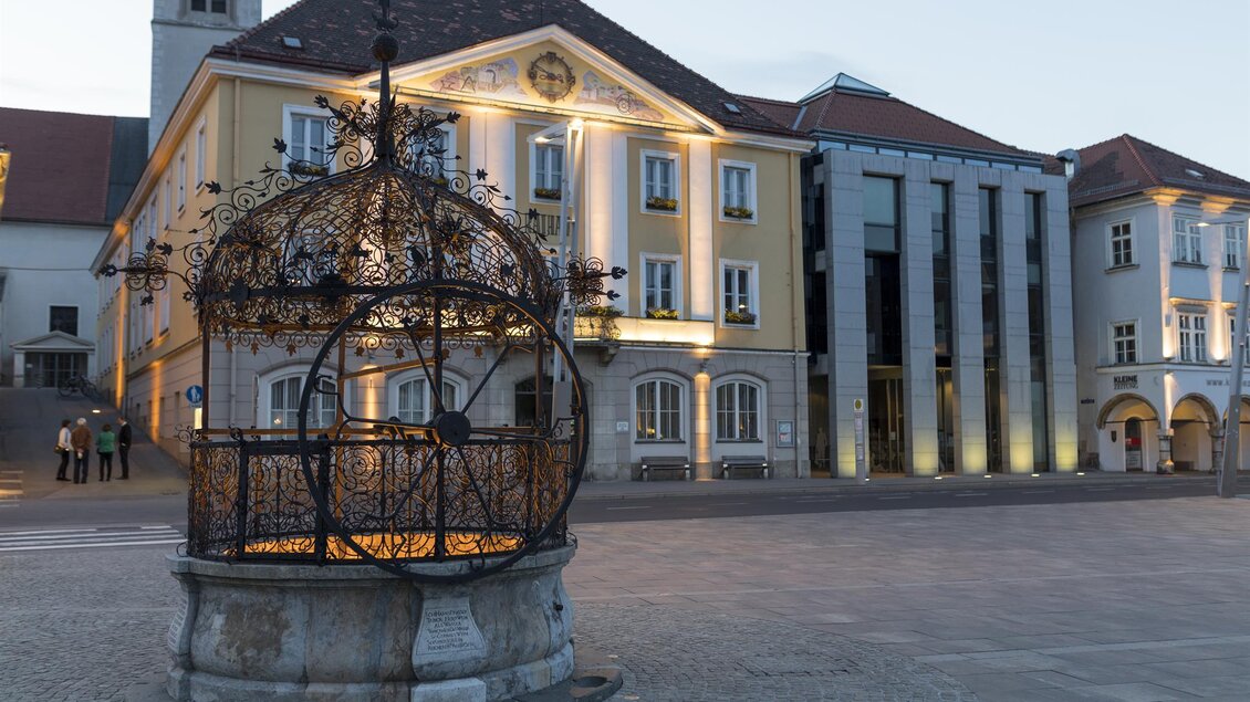 Ein historischer Brunnen mit kunstvollem Gitterwerk steht auf einem Platz. Im Hintergrund sind moderne und historische Gebäude beleuchtet. | © Martin Meieregger