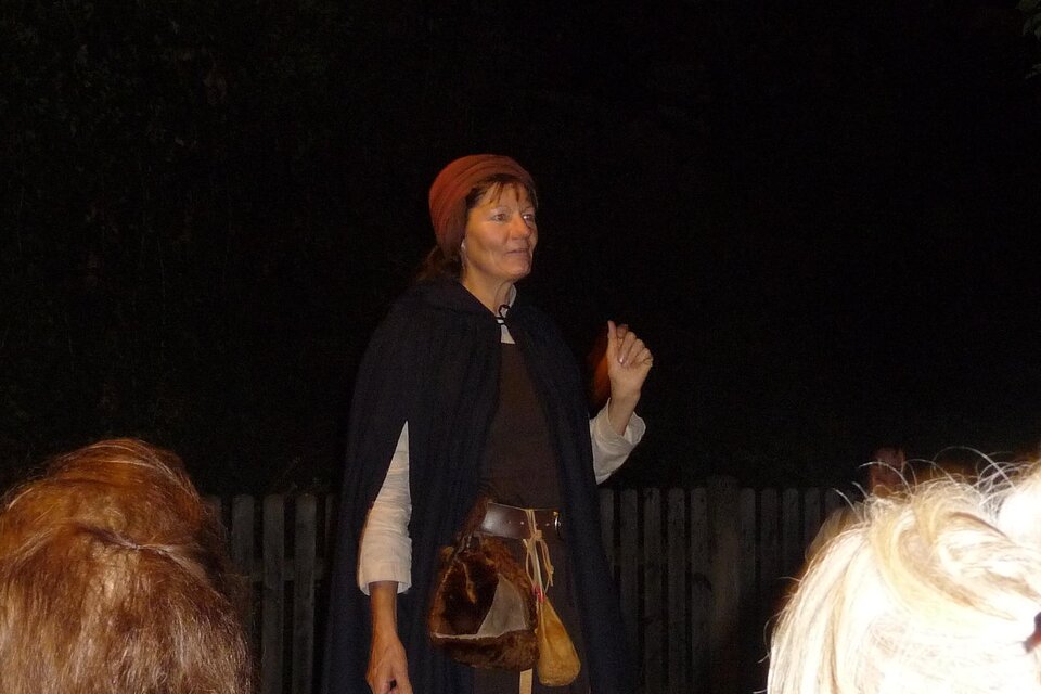 Eine Frau in historischer Kleidung steht im Dunkeln und hält eine Hand hoch. Sie trägt einen Umhang und eine Mütze, während sie anscheinend eine Geschichte erzählt. | © Mag. Helga Papst