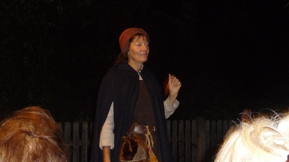 Eine Frau in historischer Kleidung steht im Dunkeln und hält eine Hand hoch. Sie trägt einen Umhang und eine Mütze, während sie anscheinend eine Geschichte erzählt. | © Mag. Helga Papst