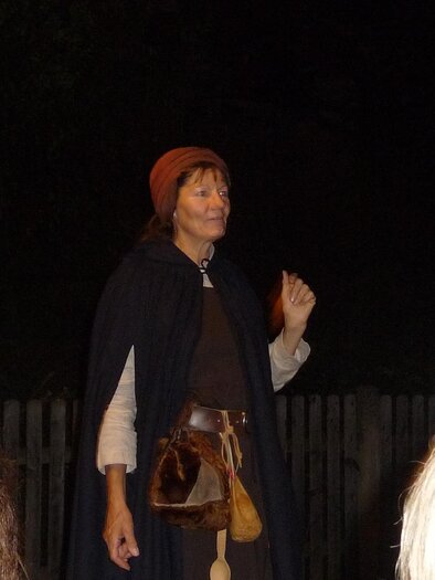 Eine Frau in historischer Kleidung steht im Dunkeln und hält eine Hand hoch. Sie trägt einen Umhang und eine Mütze, während sie anscheinend eine Geschichte erzählt. | © Mag. Helga Papst