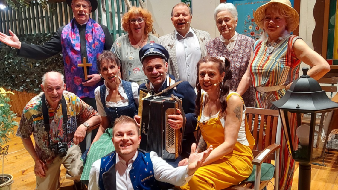Eine Gruppe von Menschen posiert fröhlich auf einer Bühne mit einer ländlichen Kulisse. Sie tragen bunte Kleidung und zeigen gute Laune. | © GANZER
