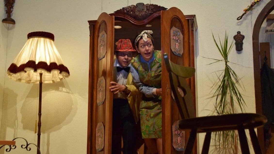 Zwei Personen verstecken sich in einem Schrank und sehen überrascht aus. Im Hintergrund sind eine Lampe und eine Pflanze zu sehen. | © Murtaler Bauernbühne