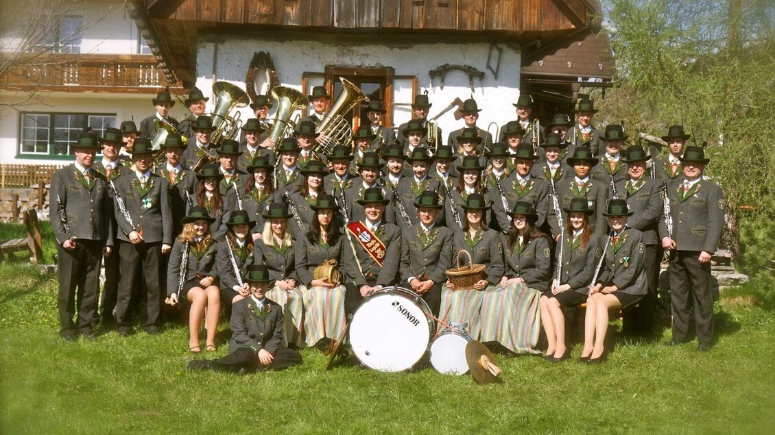 Eine große Gruppe von Menschen in traditionellen Kleidern steht vor einem Holzhaus. Sie tragen Hüte und halten Musikinstrumente, während sie fröhlich posieren. | © Musikkapelle Kumitz