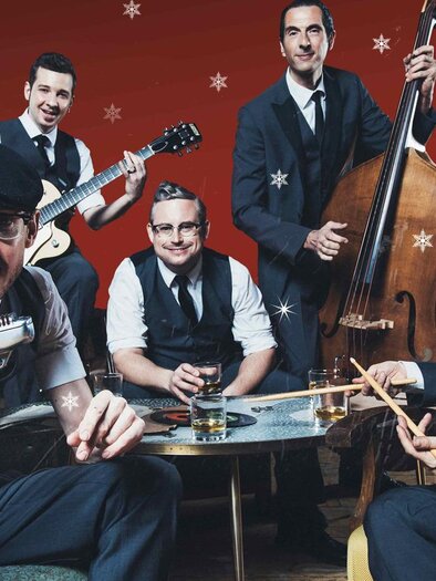 Eine Band mit fünf Musikern in eleganter Kleidung sitzt um einen Tisch. Der Hintergrund ist rot mit weißen Schneeflocken. | © Marija-Kanizaj
