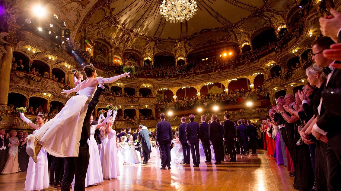 Ein festlicher Ball in einem prächtigen Saal mit einem großen Kronleuchter. Elegante Paare tanzen auf dem Parkett, umgeben von fröhlichen Gästen. | © Marija Kanizaj