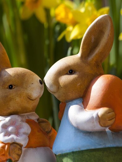 Ein Paar entzückender Osterhasenfiguren steht vor gelben Narzissen. Die Hasen sind fröhlich und tragen farbenfrohe Kleidung. | © pixabay.com