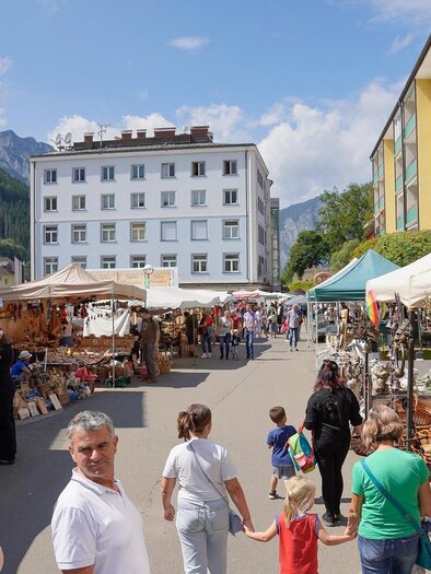 A bustling market with many stalls and visitors. In the background, mountains and several buildings can be seen. | © Christian Berger
