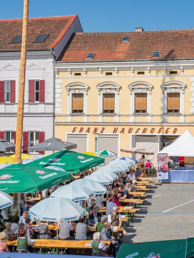 Ein belebter Marktplatz mit vielen Menschen und bunten Schirmen. Im Hintergrund sind historische Gebäude und ein strahlend blauer Himmel zu sehen. | © Marktgemeinde Pöllau