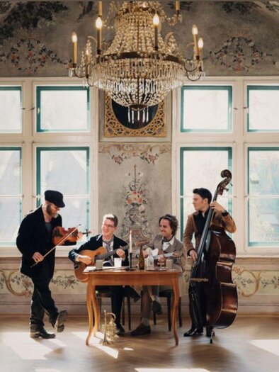 Eine musikalische Gruppe spielt in einem eleganten Raum mit einem Kronleuchter. Sie sitzen an einem Tisch und haben verschiedene Instrumente dabei. | © Kammerhofmuseum