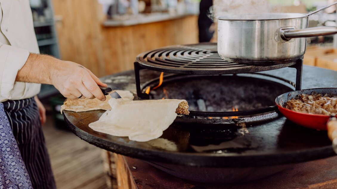 Eine Person kocht an einem offenen Grill. In der Pfanne befinden sich leckere Speisen und Dampf steigt auf. | © Karin Bergmann
