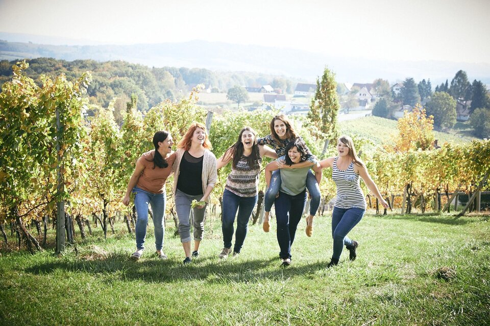 Sechs Frauen lachen und laufen fröhlich durch einen Weinberg. Im Hintergrund sind sanfte Hügel und ein sonniger Himmel zu sehen. | © Die Weinblüten-Winzerinnen im Vulkanland