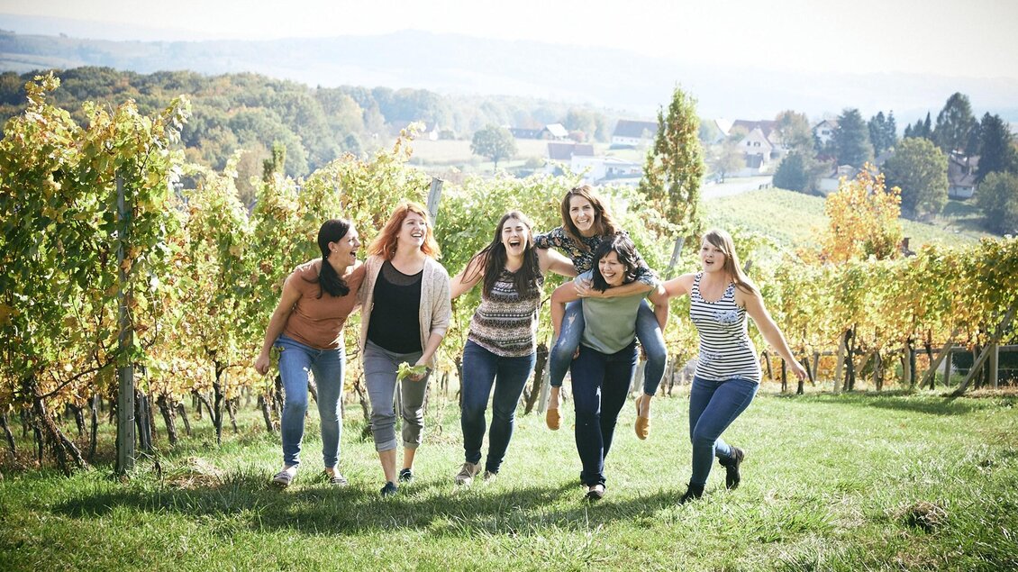 Sechs Frauen lachen und laufen fröhlich durch einen Weinberg. Im Hintergrund sind sanfte Hügel und ein sonniger Himmel zu sehen. | © Die Weinblüten-Winzerinnen im Vulkanland