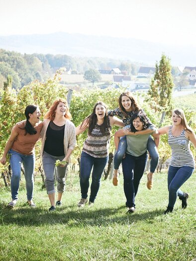Sechs Frauen lachen und laufen fröhlich durch einen Weinberg. Im Hintergrund sind sanfte Hügel und ein sonniger Himmel zu sehen. | © Die Weinblüten-Winzerinnen im Vulkanland