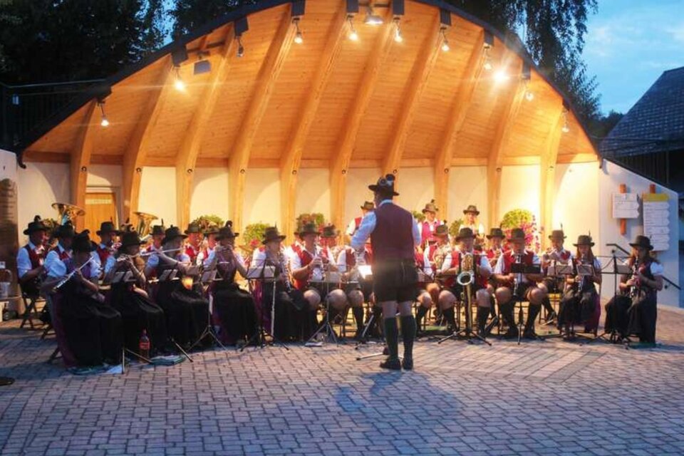 Eine große Blaskapelle spielt unter einem überdachten Bereich. Die Musiker tragen traditionelle Trachten und sind auf einem Platz im Freien versammelt. | © Wolfgang Sölkner