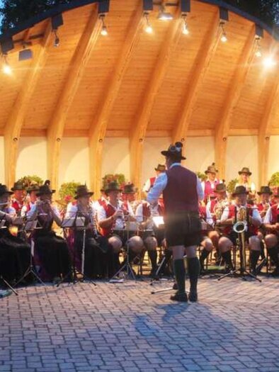 A large brass band is playing under a covered area. The musicians are wearing traditional costumes and are gathered in an outdoor square. | © Wolfgang Sölkner