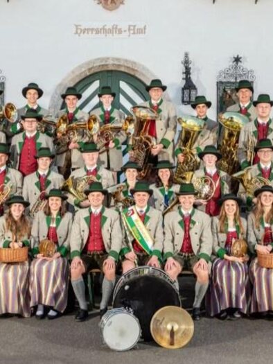 A music society performs on a stage, surrounded by festive decorations. The musicians wear green uniforms and play various instruments. | © Musikverein Öblarn