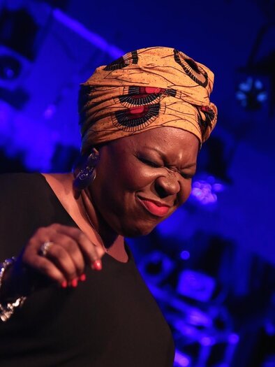 A singer with a colorful headscarf stands on stage and sings passionately. In the background, soft lights create a cheerful atmosphere. | ©  Aleksandra Pruenner