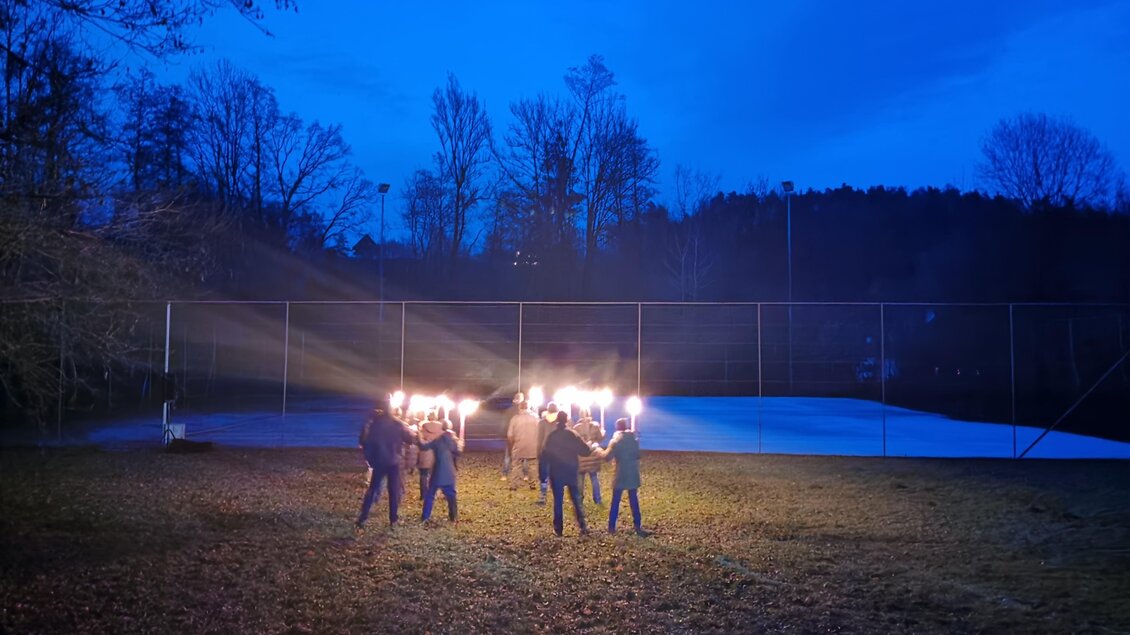 Eine Gruppe von Menschen steht im Freien und hält Fackeln in der Dämmerung. Im Hintergrund sind Bäume und ein Sportplatz zu sehen. | © CDM