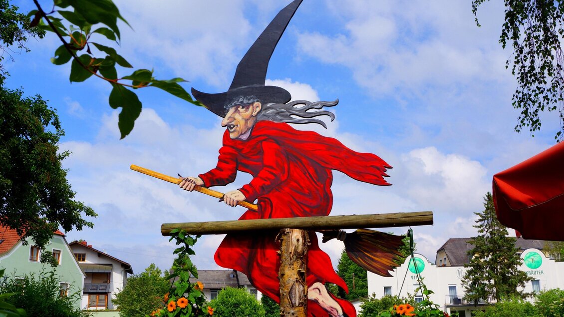 Eine große, bunte Hexenfigur fliegt auf einem Besen durch den Himmel. Im Vordergrund sind Blumen und grüne Blätter zu sehen. | © Manuela Schweinzer-Riegler