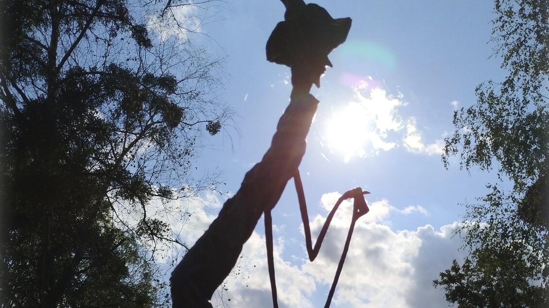 Eine silhouette eines Kunstwerks zeigt eine Figur mit Hut, die eine Pose einnimmt. Im Hintergrund strahlt die Sonne zwischen den Wolken. | © Manuela Schweinzer-Riegler