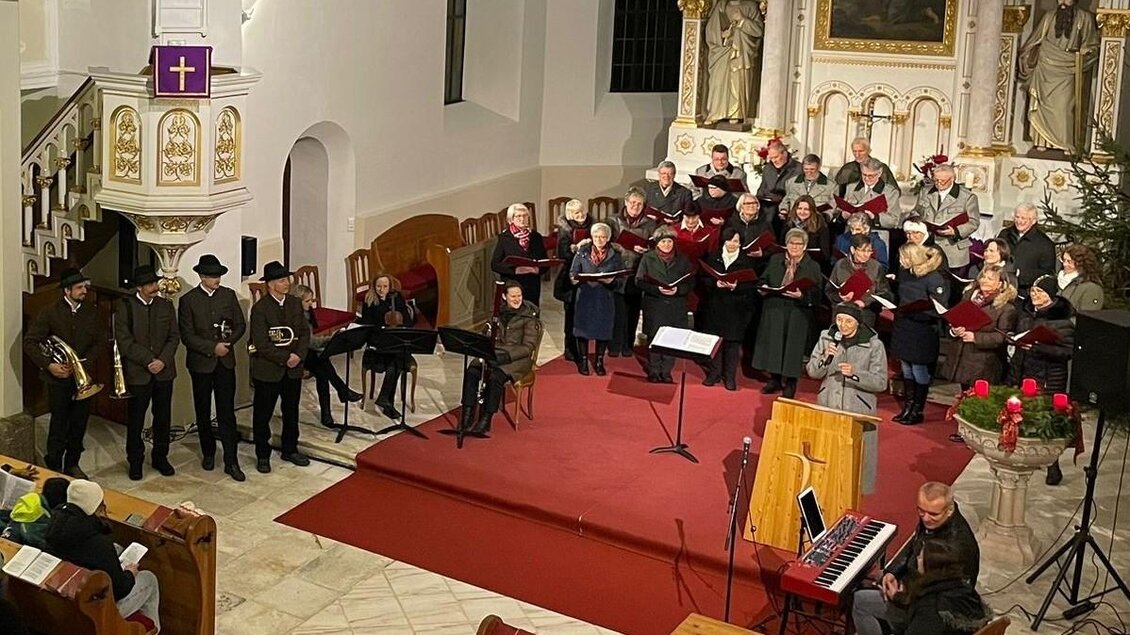 Eine Choraufführung in einer Kirche mit vielen Sängern, die in festlicher Kleidung da stehen. Im Vordergrund sind Musiker und Zuschauer zu sehen, während die Atmosphäre festlich und einladend ist. | © Hirnbirnmusi