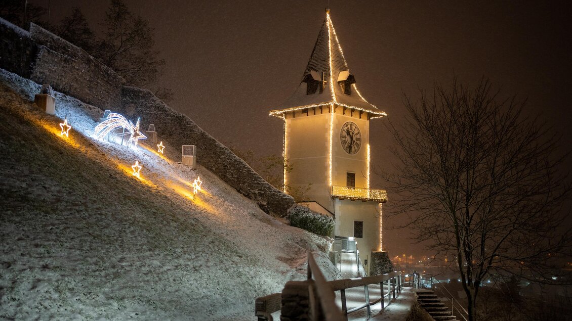 Eine verschneite Landschaft mit einem beleuchteten Turm. Im Hintergrund sind Weihnachtslichter und der Nachthimmel zu sehen. | © TV Hochsteiermark / Ekatarina Paller