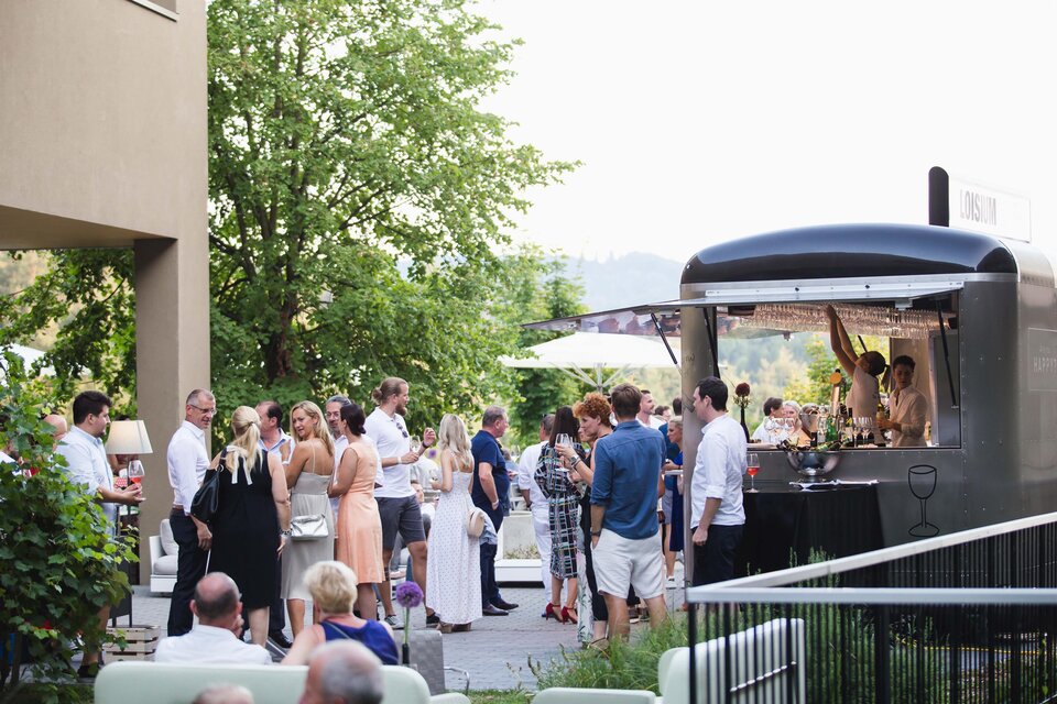 Eine fröhliche Menschenmenge steht an einem Food Truck und genießt den Abend. Im Hintergrund sind Bäume und ein klarer Himmel zu sehen. | © LOISIUM