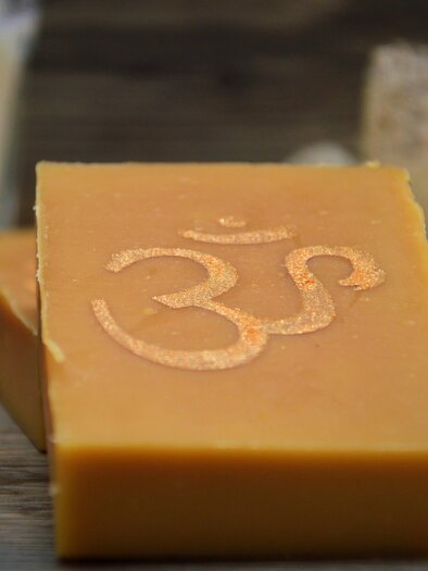 Eine Seife mit dem Symbol „Om“ auf der Oberfläche liegt auf einem Holzuntergrund. Im Hintergrund sind einige natürliche Materialien und eine Flasche zu sehen. | © Martina Schmidt