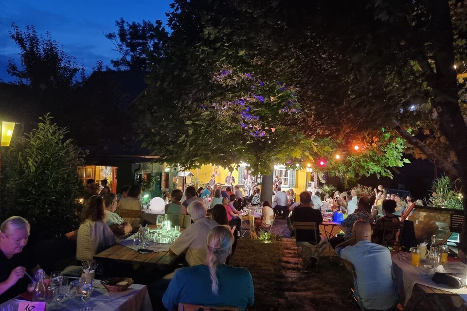 Ein gemütlicher Außenbereich mit Tischreihen unter einem Baum, beleuchtet von bunten Lichtern. Menschen genießen den Abend in einer geselligen Atmosphäre. | © Kürzl