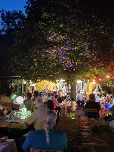 Ein gemütlicher Außenbereich mit Tischreihen unter einem Baum, beleuchtet von bunten Lichtern. Menschen genießen den Abend in einer geselligen Atmosphäre. | © Kürzl