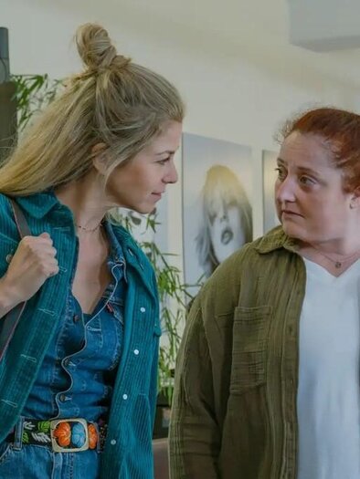 Two women are standing in a modern room and having a conversation. One is wearing a green jacket and jeans, while the other is wearing a white top and a khaki jacket. | © Oststeiermark Tourismus