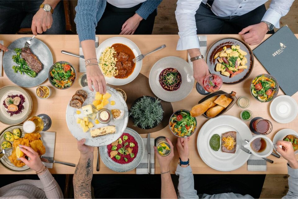 Ein schöner Tisch voller köstlicher Gerichte, darunter Fleisch, Salate und verschiedene Beilagen. Mehrere Personen genießen gemeinsam das Essen und probieren die Leckereien. | © MONDI Resort Grundlsee