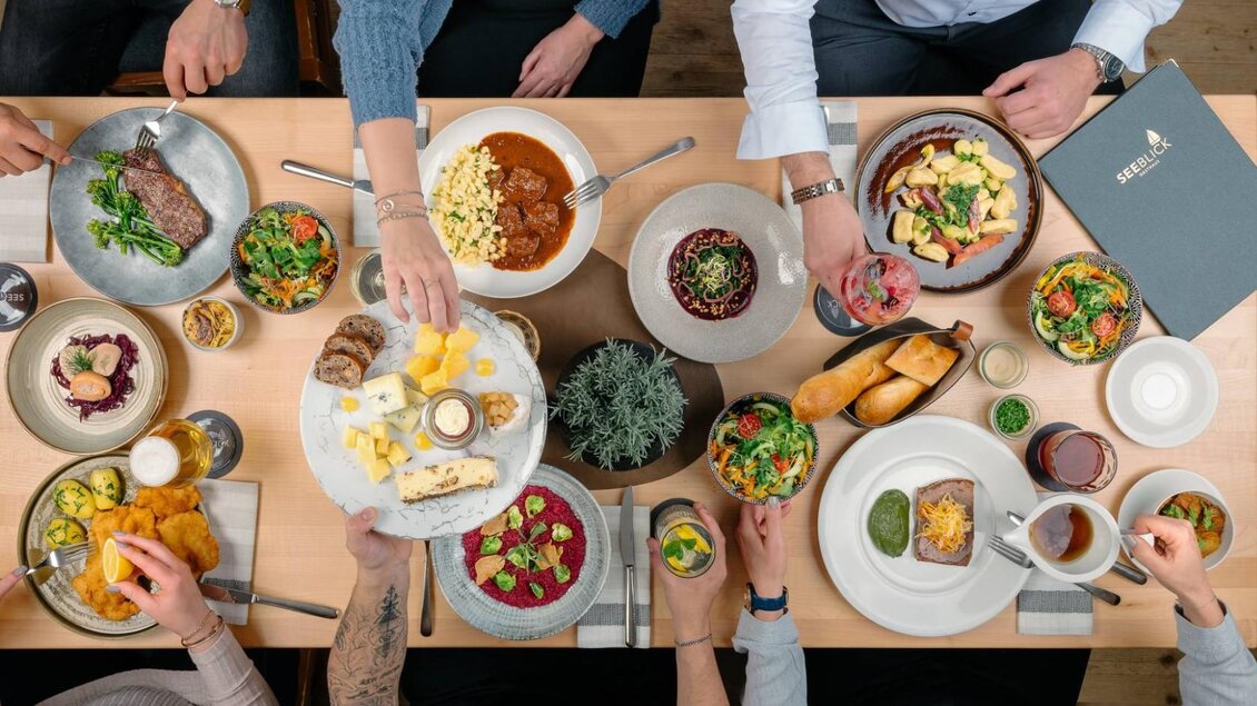 Ein schöner Tisch voller köstlicher Gerichte, darunter Fleisch, Salate und verschiedene Beilagen. Mehrere Personen genießen gemeinsam das Essen und probieren die Leckereien. | © MONDI Resort Grundlsee