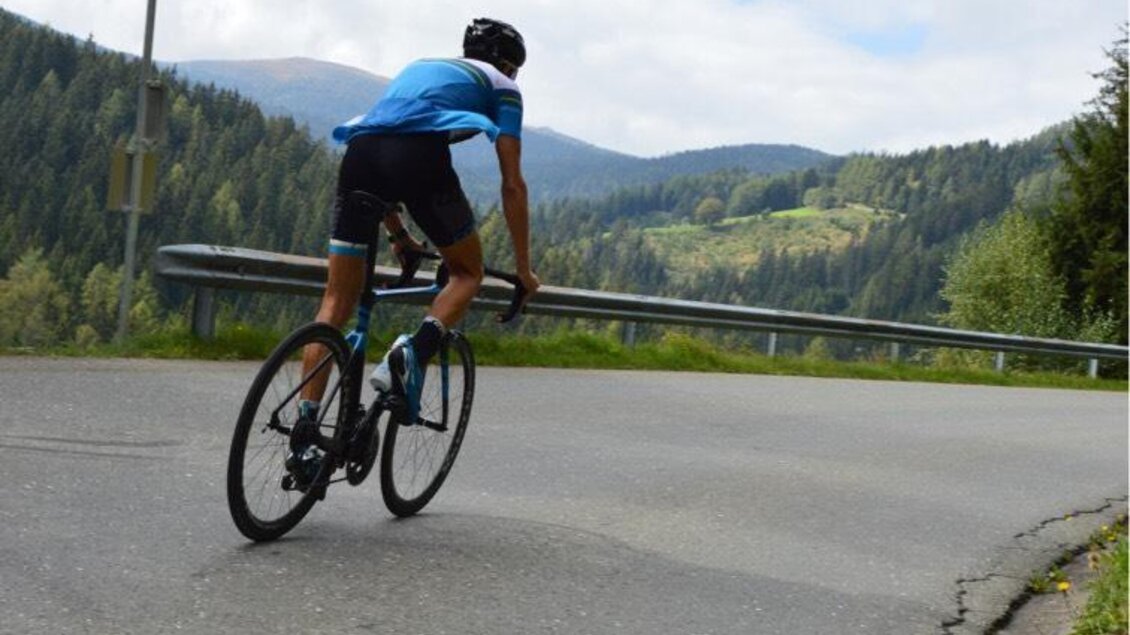 Ein Radfahrer fuhrt einen Hügel hinauf auf einer ländlichen Straße. Im Hintergrund sind grüne Berge und ein bewölkter Himmel zu sehen. | © EvenDZ