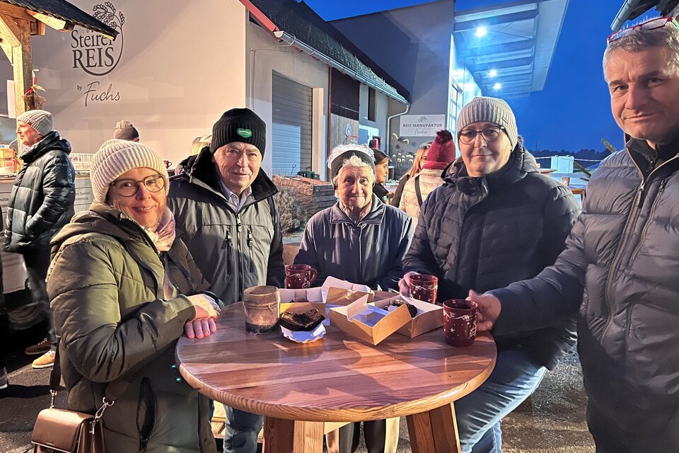 Eine Gruppe von fünf Personen steht um einen runden Holztisch. Sie tragen warme Kleidung und genießen Getränke in einer gemütlichen Umgebung. | © Fuchs Adele