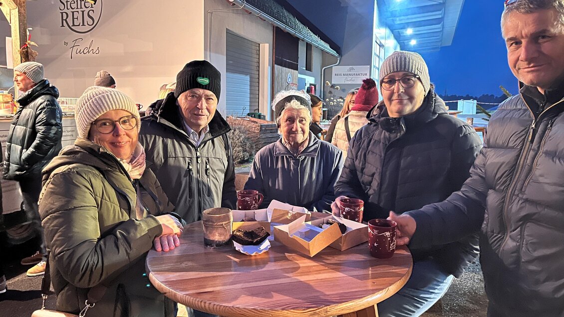 Eine Gruppe von fünf Personen steht um einen runden Holztisch. Sie tragen warme Kleidung und genießen Getränke in einer gemütlichen Umgebung. | © Fuchs Adele