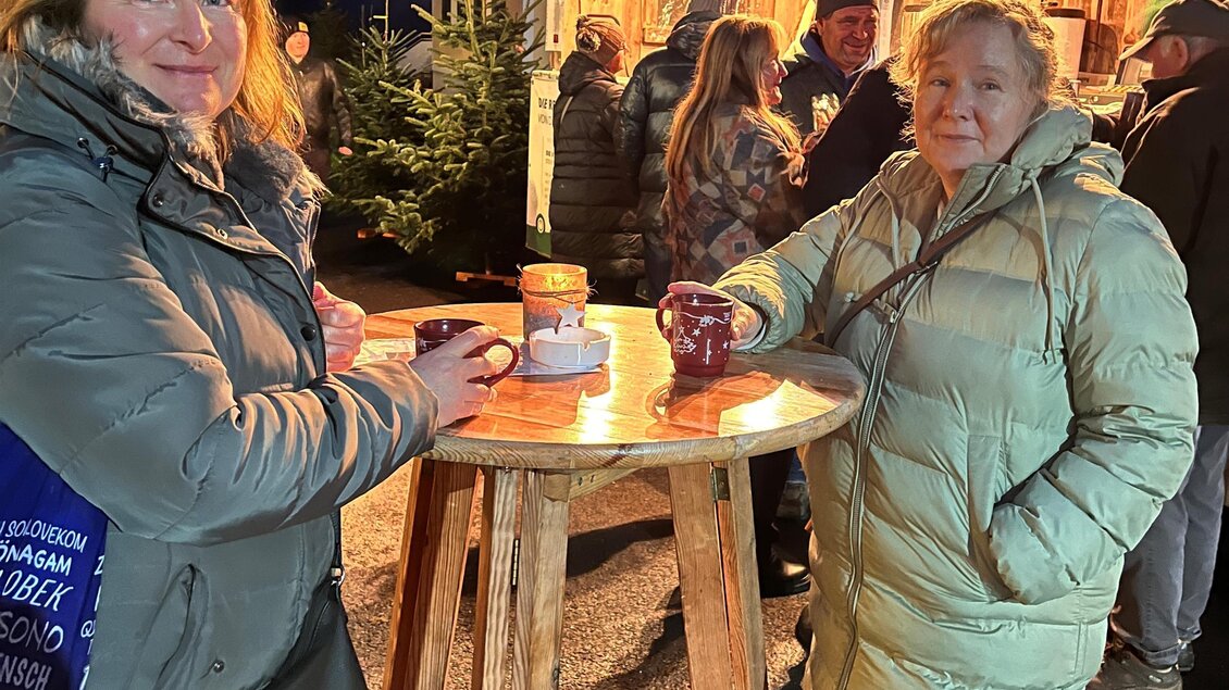 Zwei Frauen stehen an einem Stehtisch und halten Getränke in der Hand. Im Hintergrund sind weitere Personen und eine festliche Beleuchtung zu sehen. | © Fuchs Adele