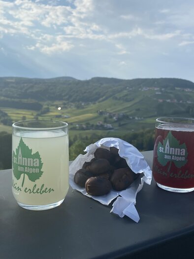 Zwei Gläser mit Getränken stehen auf einem Tisch, links ein klares, weißes Getränk und rechts ein rotes Getränk. Neben den Gläsern liegt eine Packung mit Snacks und im Hintergrund erstreckt sich eine grüne Landschaft. | © Karin Grießbacher