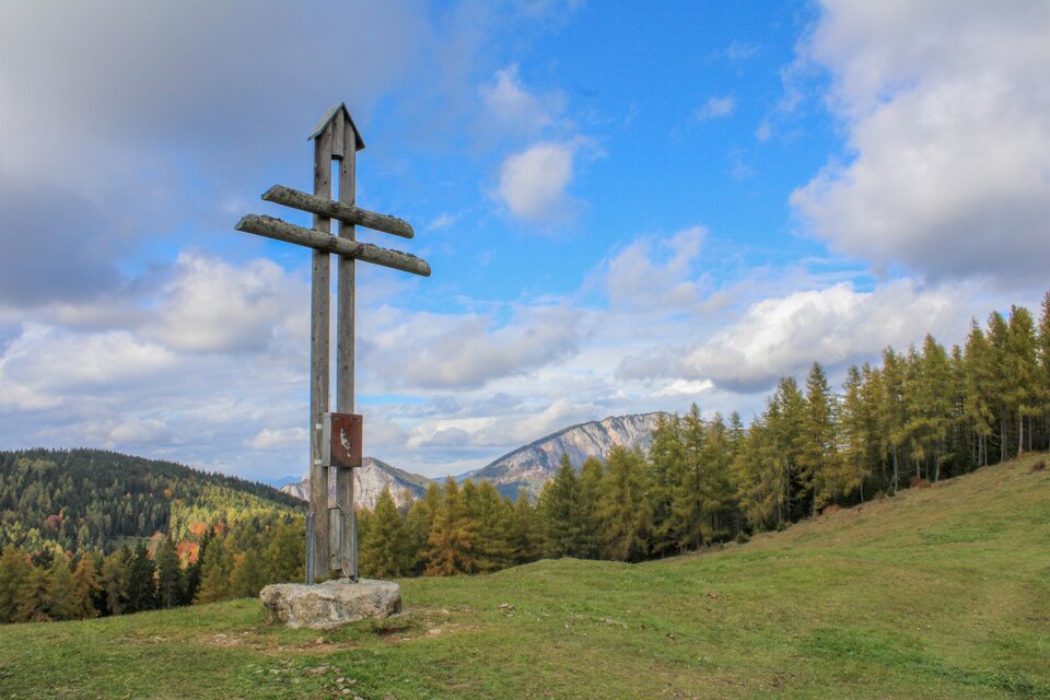 Ein großes Holzkreuz steht auf einer grünen Wiese. Im Hintergrund sind Berge und Wälder unter einem blauen Himmel zu sehen. | © Trieb Laura