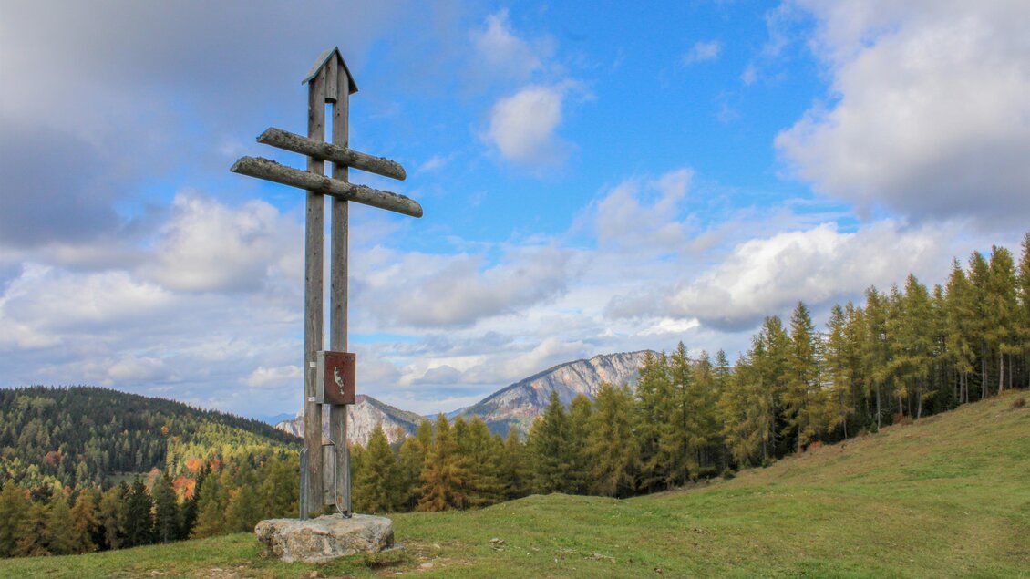Ein großes Holzkreuz steht auf einer grünen Wiese. Im Hintergrund sind Berge und Wälder unter einem blauen Himmel zu sehen. | © Trieb Laura