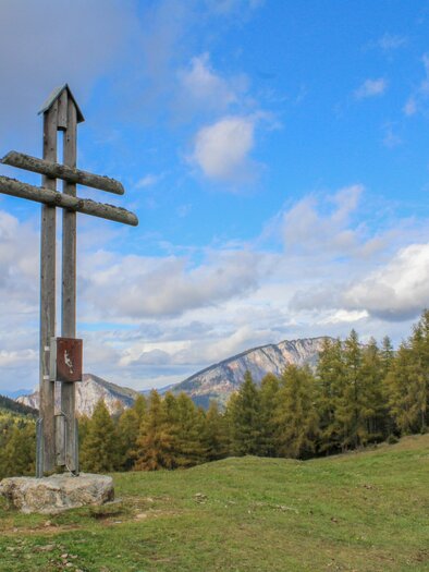 A large wooden cross stands on a green meadow. In the background, mountains and forests can be seen under a blue sky. | © Trieb Laura